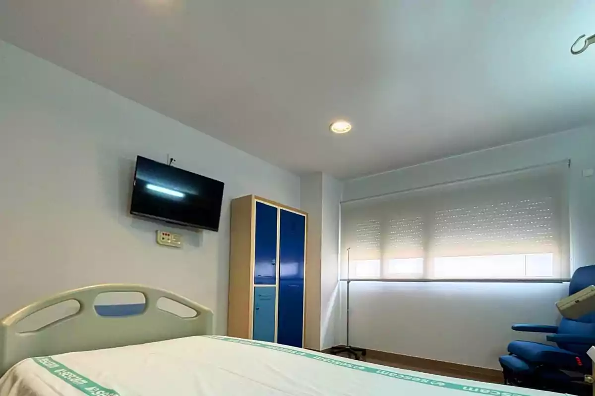 Habitación de hospital con una cama, televisor en la pared, armario azul, ventana con persiana y una silla azul. Habitación de hospital con una cama, televisor en la pared, armario azul, ventana con persiana y una silla azul.