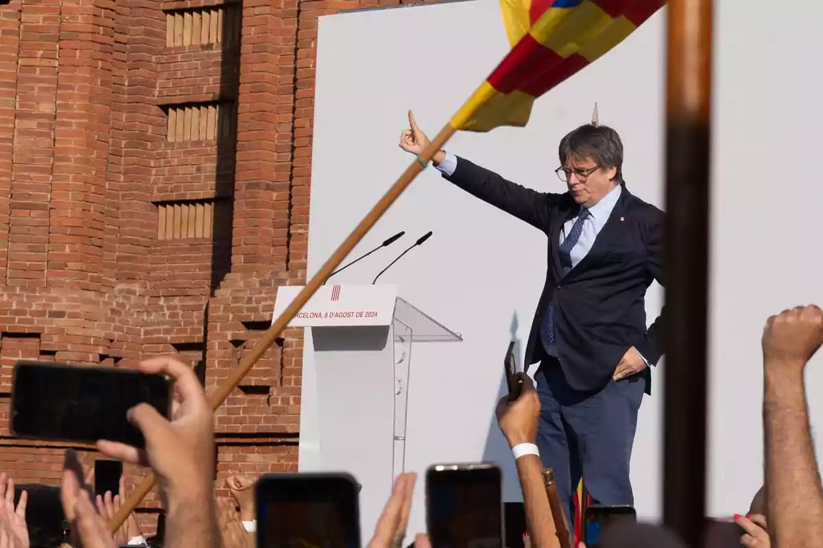 Un hombre de traje y corbata levanta el brazo en un gesto de saludo durante un evento público, con una multitud de personas sosteniendo teléfonos y banderas en el fondo, frente a un edificio de ladrillo.