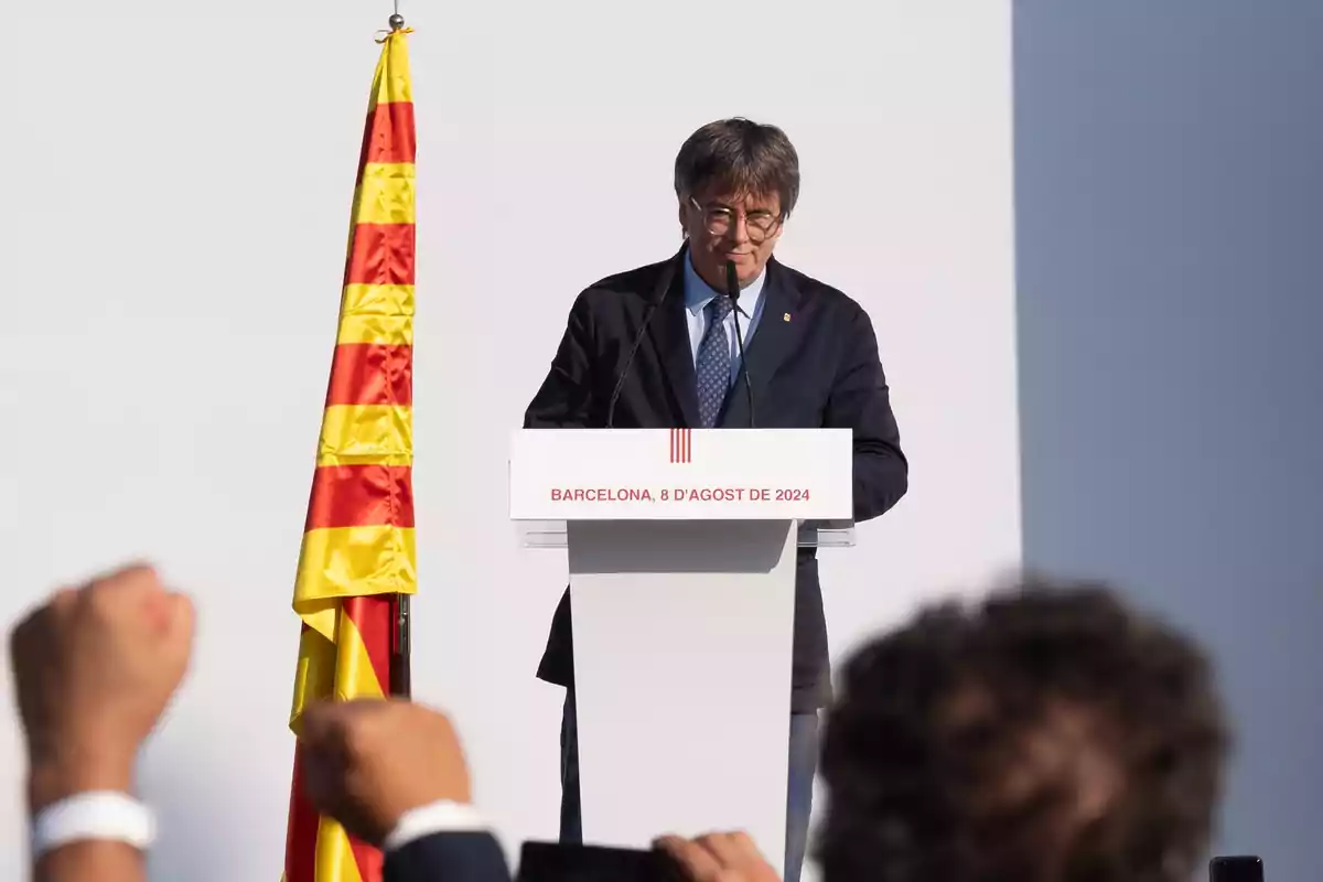 Persona dando un discurso en un podio con una bandera catalana a su lado y puños levantados en primer plano.