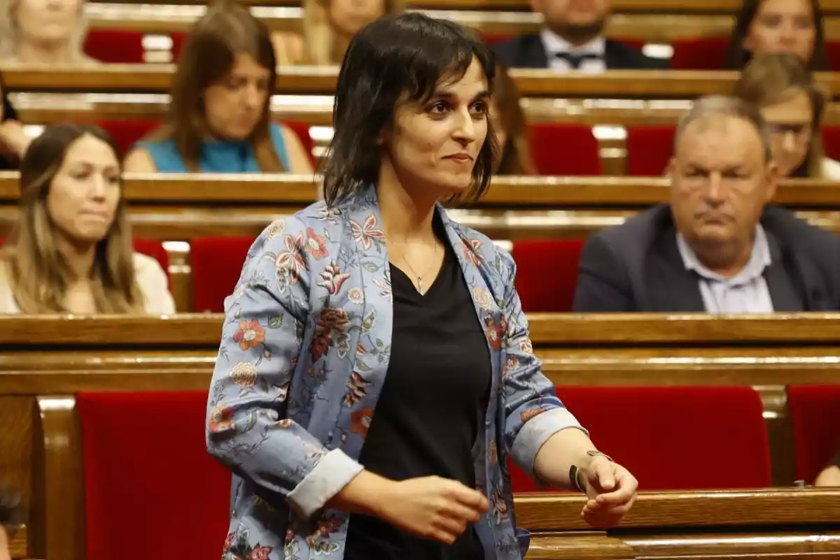 Una mujer de pie en una sala con asientos rojos y otras personas sentadas al fondo.