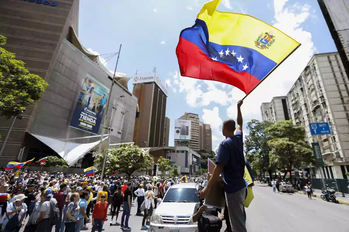 Una multitud se reúne en una calle de la ciudad mientras una persona sostiene una gran bandera de Venezuela.