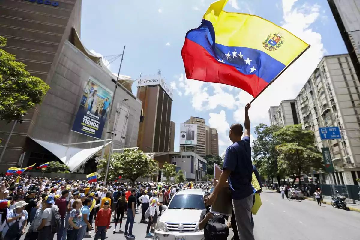 Una multitud de personas se reúne en una calle de la ciudad, mientras un hombre sostiene en alto una bandera de Venezuela.