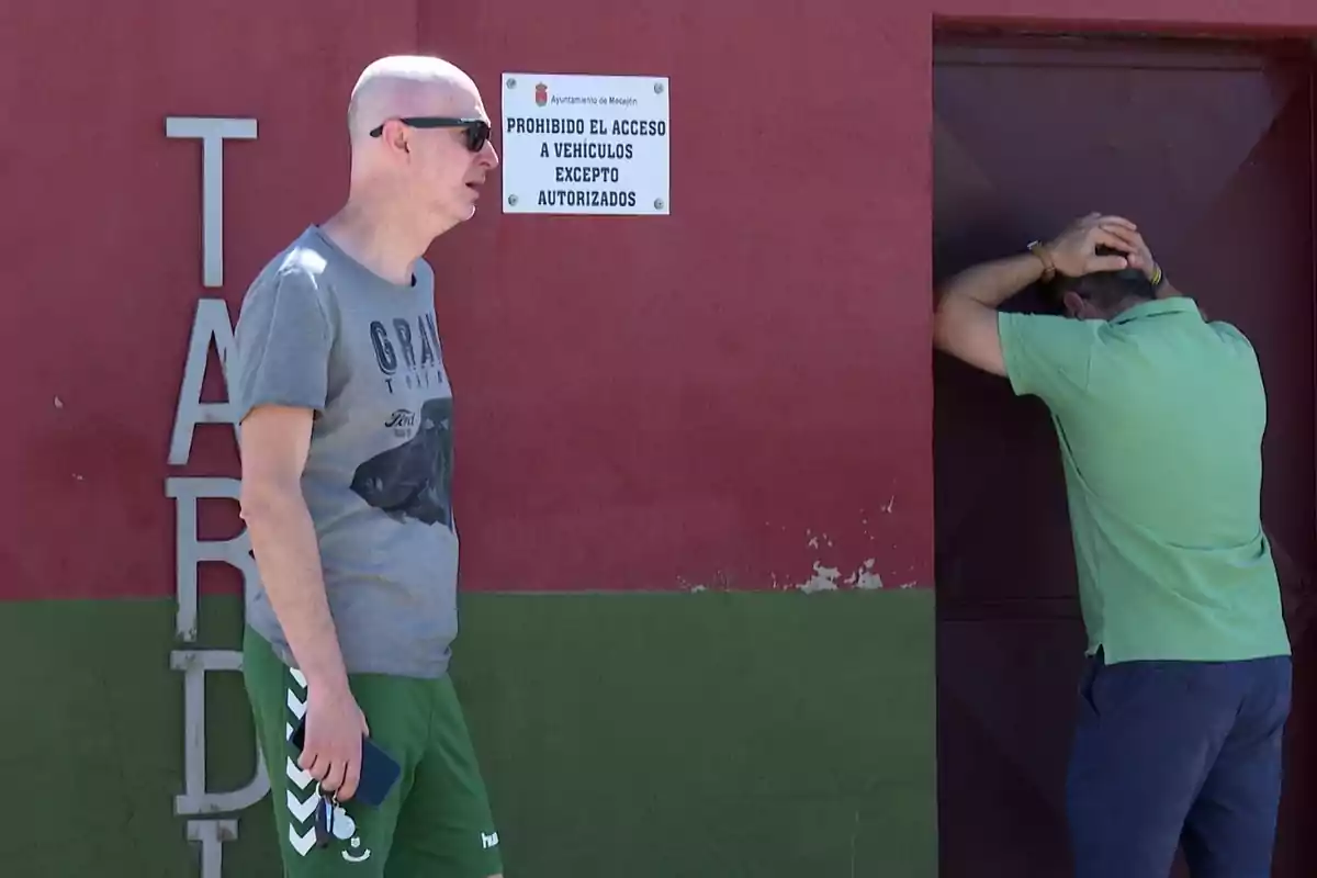 Dos hombres están frente a una pared roja y verde, uno de ellos lleva gafas de sol y ropa deportiva, mientras que el otro está apoyado en la pared con las manos en la cabeza; hay un cartel que dice "PROHIBIDO EL ACCESO A VEHÍCULOS EXCEPTO AUTORIZADOS".