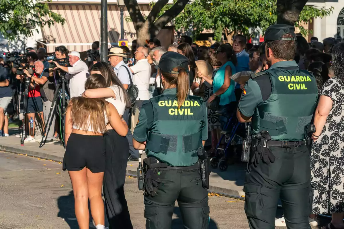 Una multitud se reúne en un evento al aire libre, con varios guardias civiles presentes y personas abrazándose mientras los medios de comunicación capturan el momento.