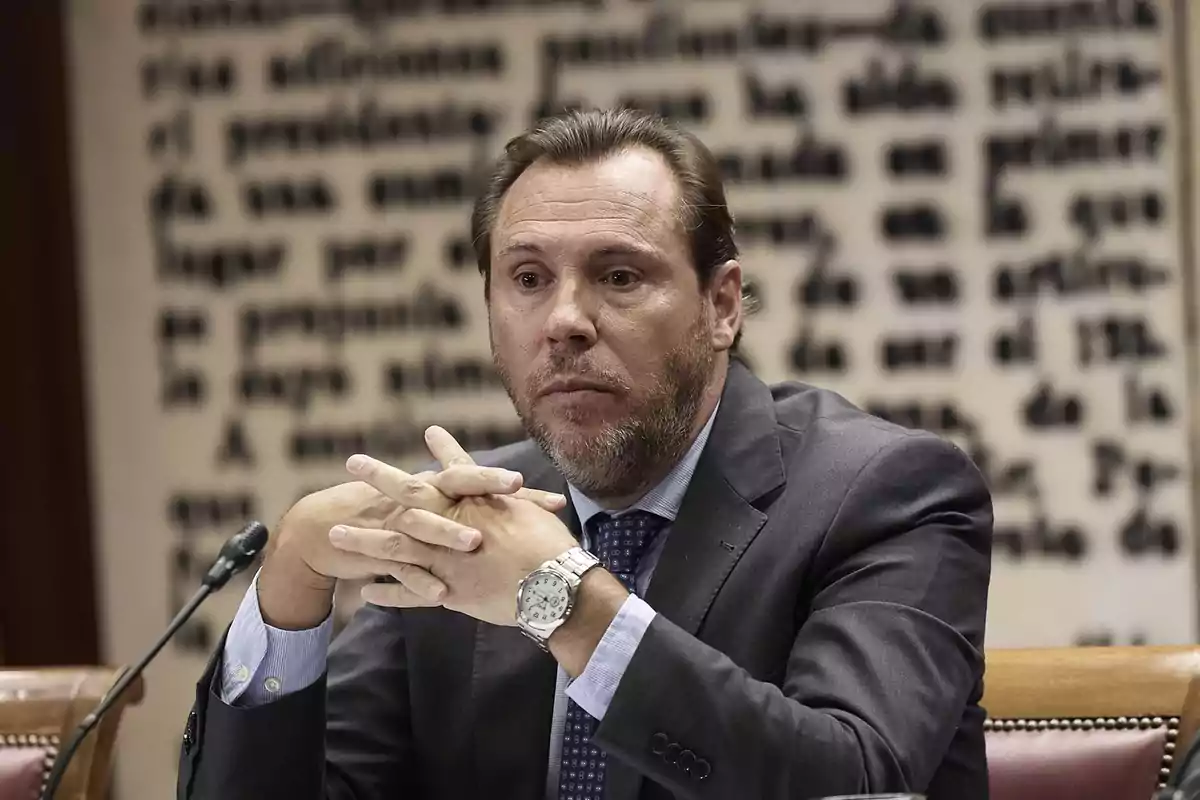 Hombre con traje y corbata sentado en una mesa con micrófono y fondo de texto borroso.