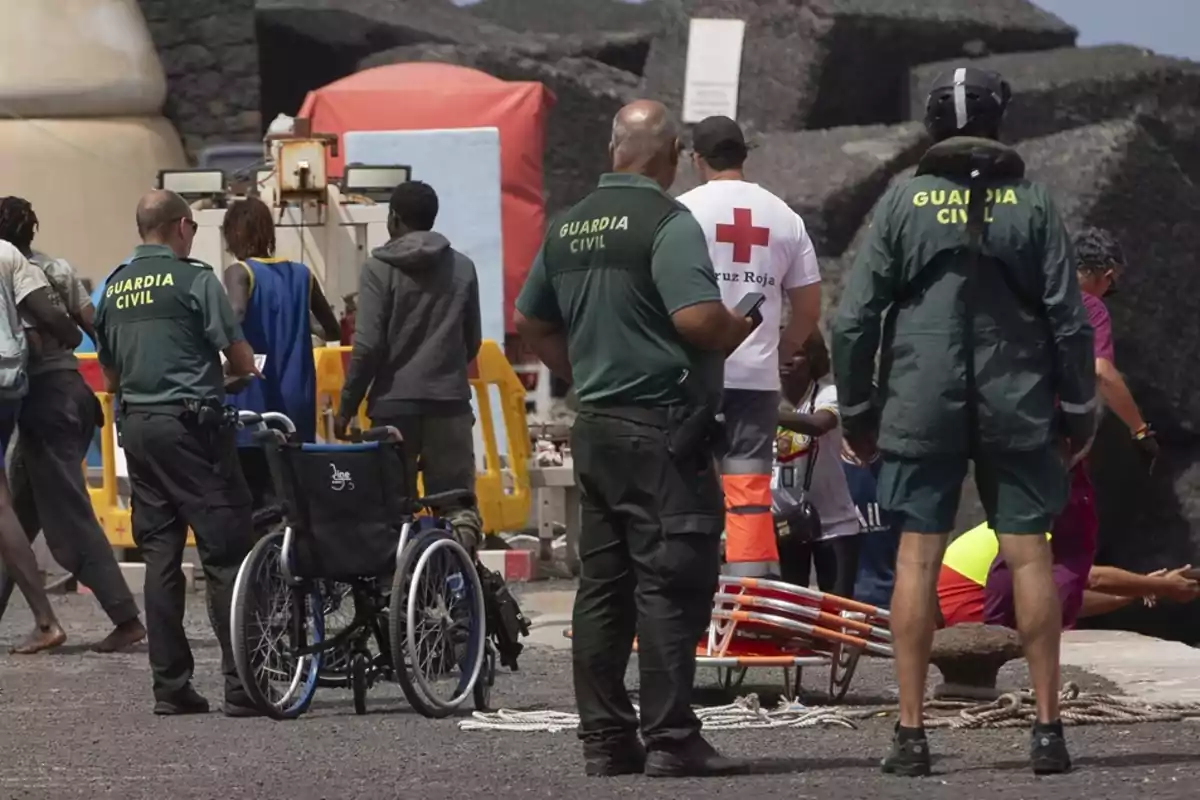 En la imagen se observa a varios miembros de la Guardia Civil y de la Cruz Roja asistiendo a un grupo de personas, algunas de las cuales parecen migrantes, en un entorno portuario.