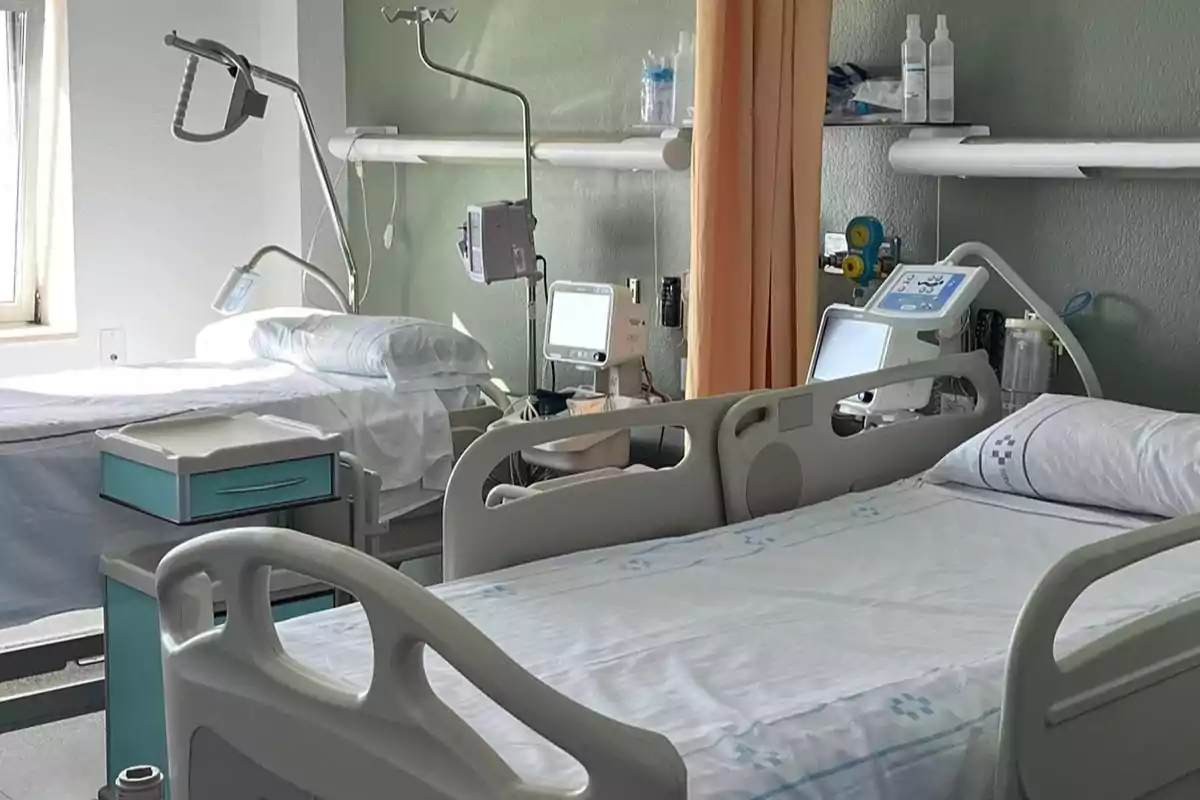 Una habitación de hospital con dos camas vacías, equipadas con monitores médicos y lámparas, junto a una ventana que deja entrar luz natural.