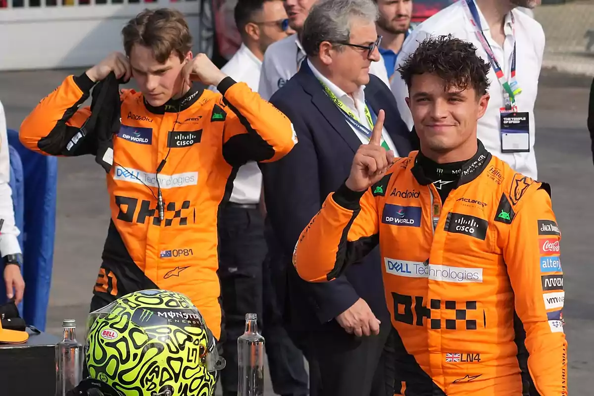 Dos pilotos de carreras con trajes naranjas, uno ajustándose el casco y el otro levantando el dedo índice, con varias personas en el fondo.