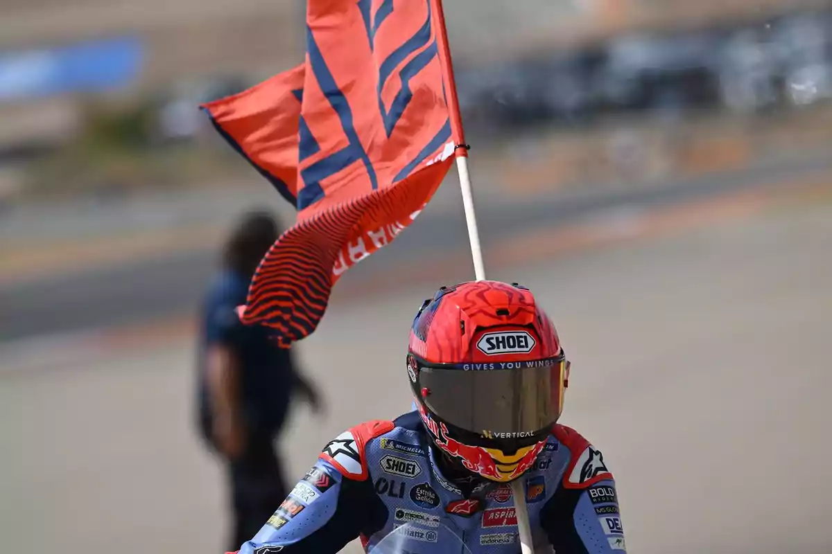 Motociclista con casco rojo y azul sosteniendo una bandera roja en una pista de carreras.