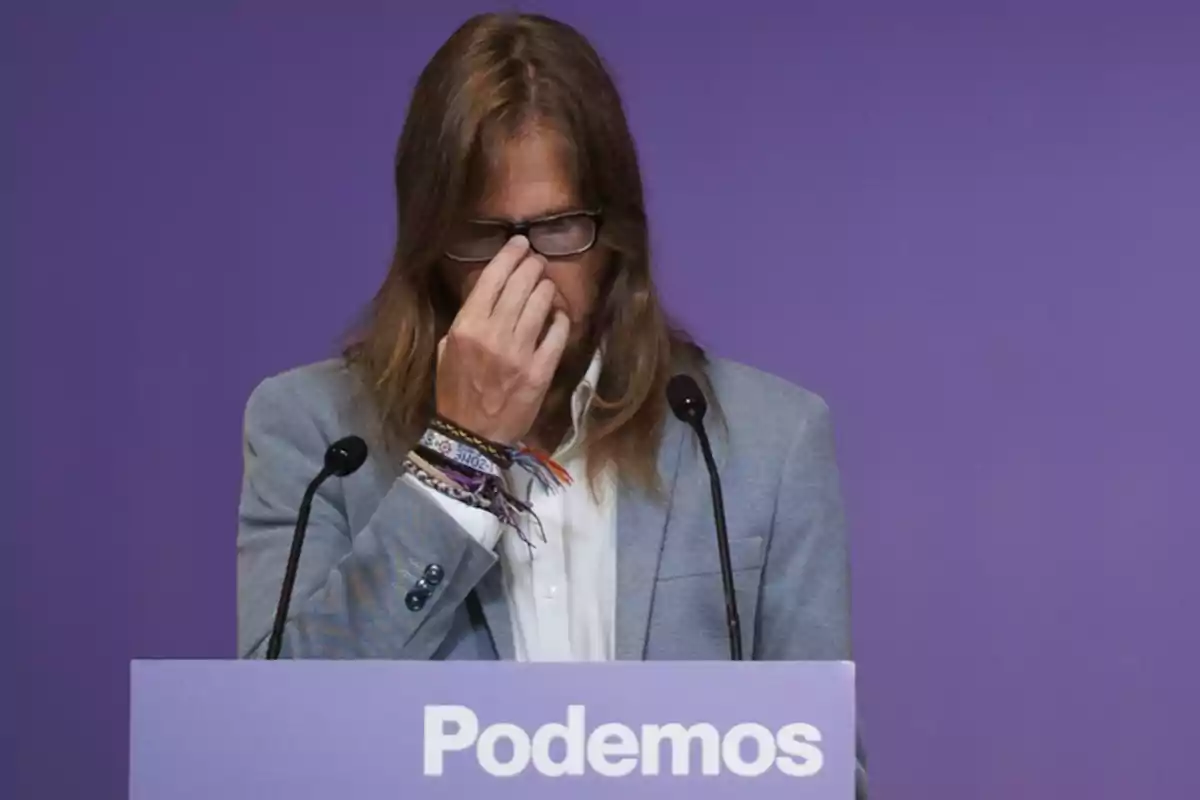 Una persona con cabello largo y gafas se encuentra detrás de un atril con la palabra "Podemos" en un fondo morado, tocándose la cara con una mano.