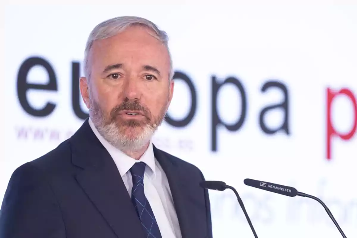 Hombre de traje y corbata hablando en un podio con un micrófono, con un fondo que tiene la palabra "europa" en letras grandes.