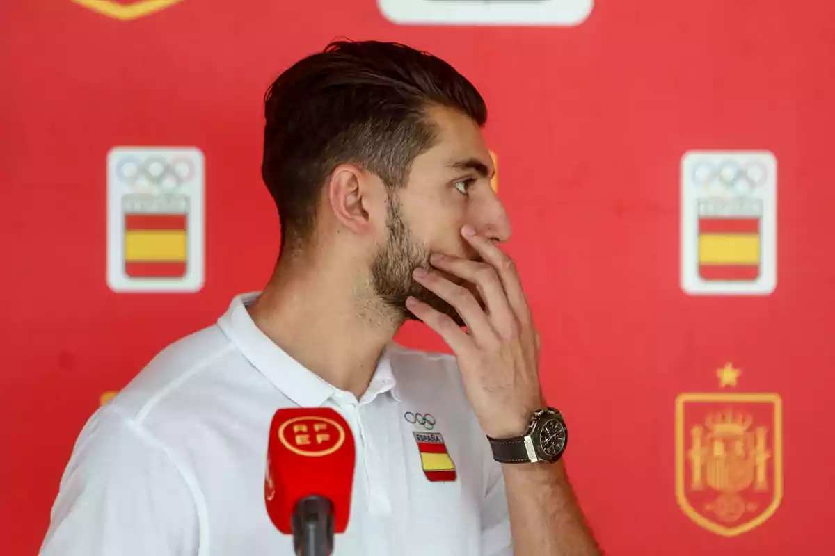 Un hombre con camiseta blanca y logotipo de España, frente a un micrófono rojo con fondo rojo y escudos olímpicos y de la federación española.