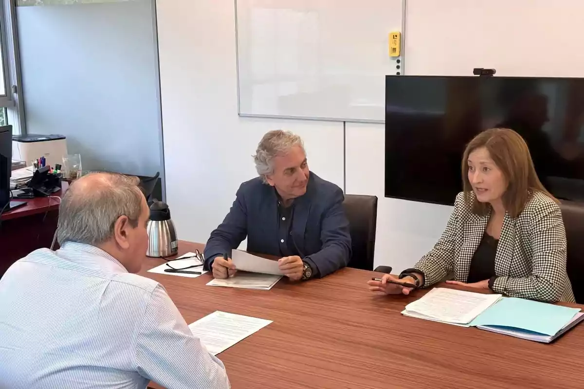 Three people are sitting around a table in an office, participating in a meeting and reviewing documents.