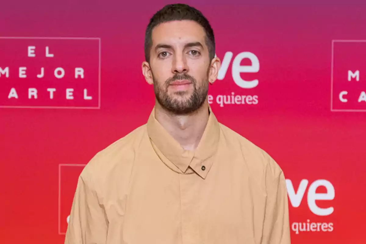 Hombre con barba y cabello corto, vestido con una camisa beige, posando frente a un fondo rojo con texto que dice "El Mejor Cartel" y "te quieres".