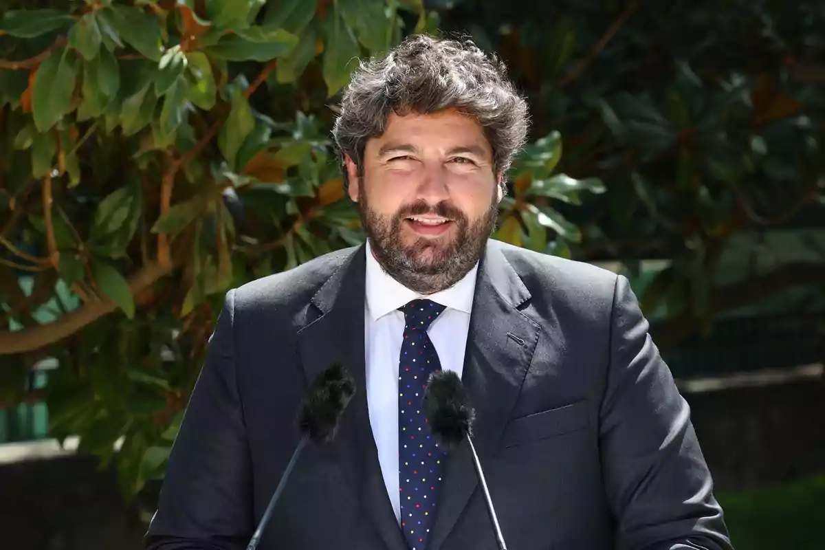 Hombre con traje y corbata hablando frente a dos micrófonos con un árbol de fondo.