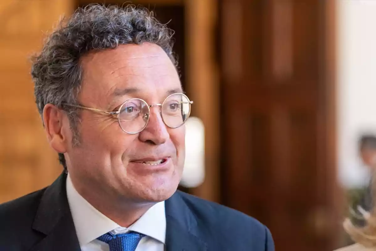 Un hombre con gafas y cabello rizado sonríe mientras lleva un traje y corbata azul en un entorno interior.