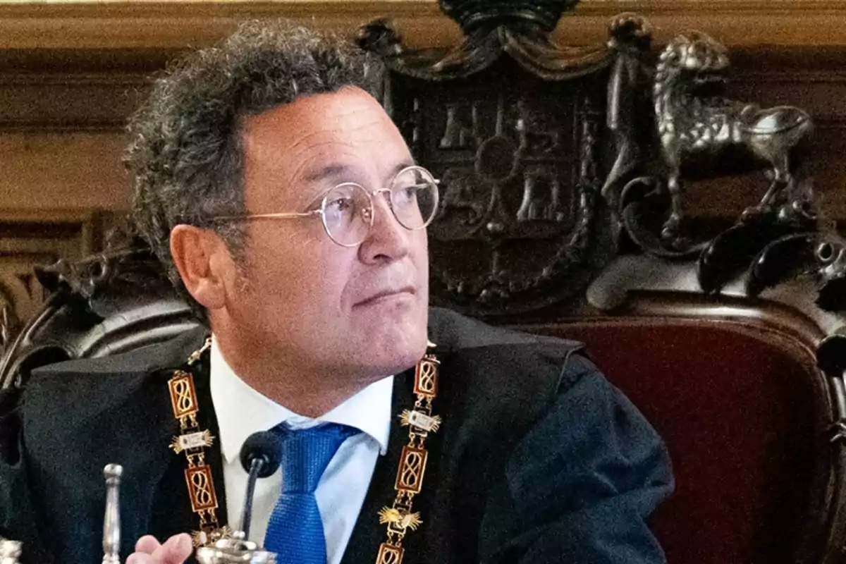 Un hombre con gafas y traje formal sentado en una silla ornamentada.