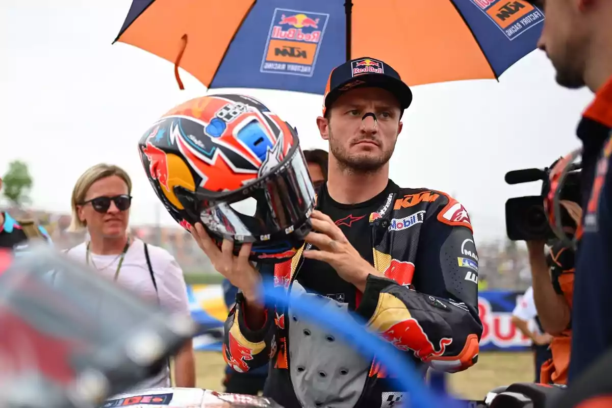 Un piloto de motociclismo sostiene su casco mientras está bajo una sombrilla de Red Bull KTM, rodeado de personas y cámaras.