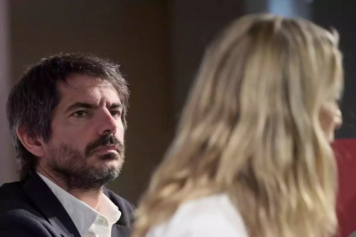 Hombre con barba y cabello oscuro mirando hacia adelante mientras una mujer rubia está desenfocada en primer plano. Hombre con barba y cabello oscuro mirando hacia adelante mientras una mujer rubia está desenfocada en primer plano.