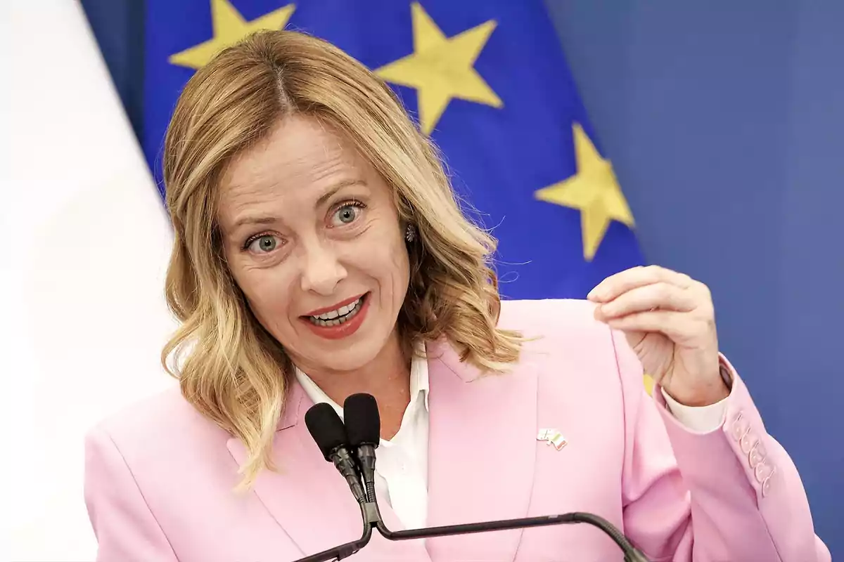 Una mujer con chaqueta rosa hablando frente a micrófonos con una bandera de la Unión Europea de fondo.