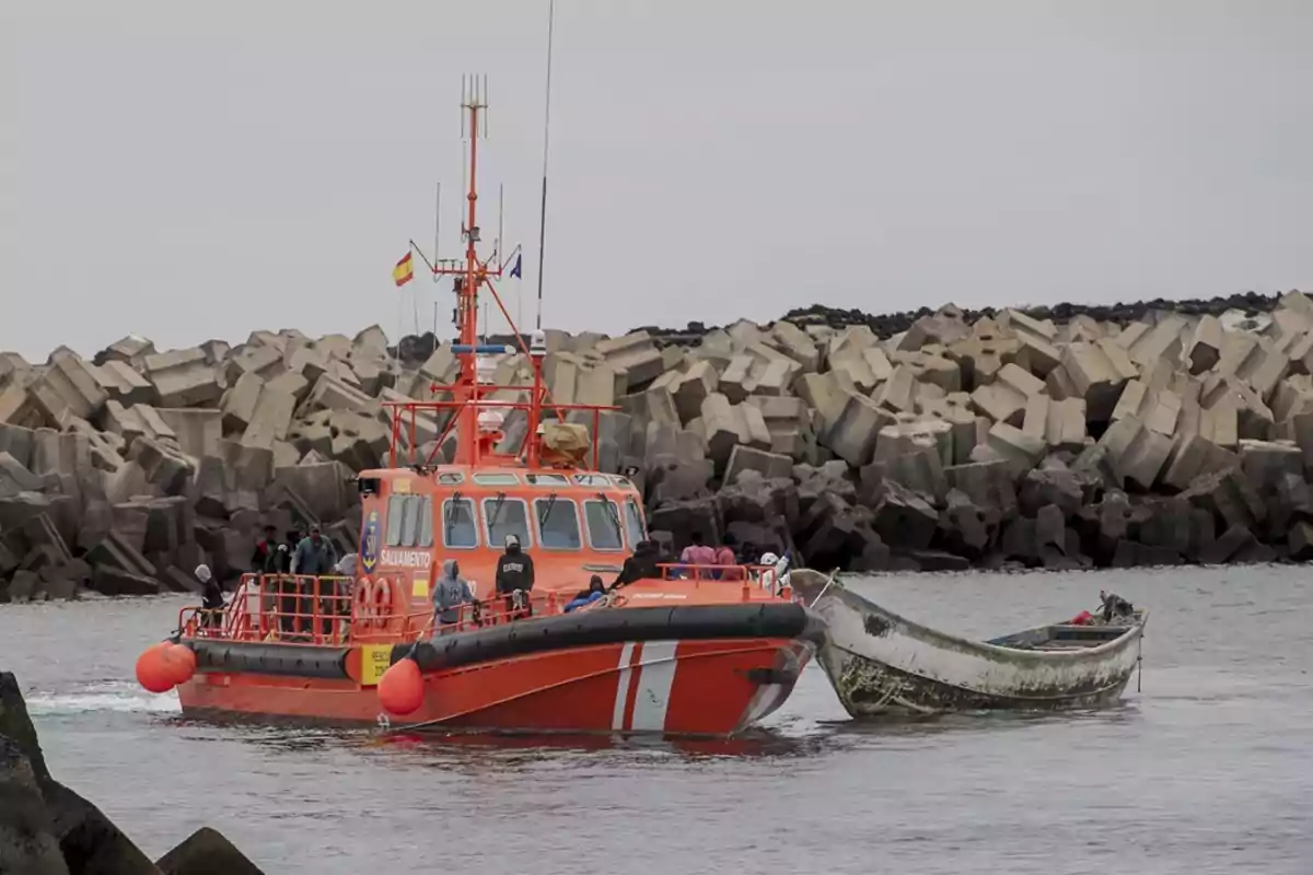 Una embarcación de rescate de color naranja remolca una lancha en el agua cerca de un rompeolas de bloques de hormigón.