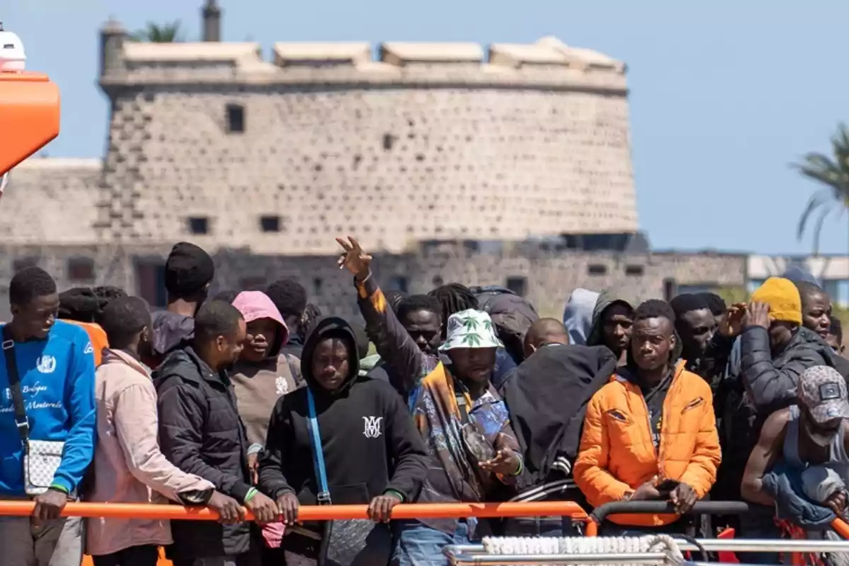 Un grupo de personas, algunas con ropa colorida y abrigos, se encuentran reunidas al aire libre frente a una estructura de piedra.