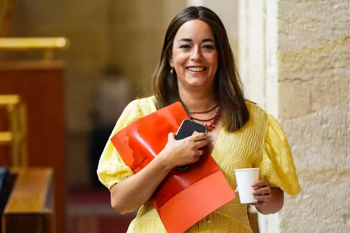 Mujer sonriente con vestido amarillo sosteniendo una carpeta roja, un teléfono móvil y un vaso de papel.