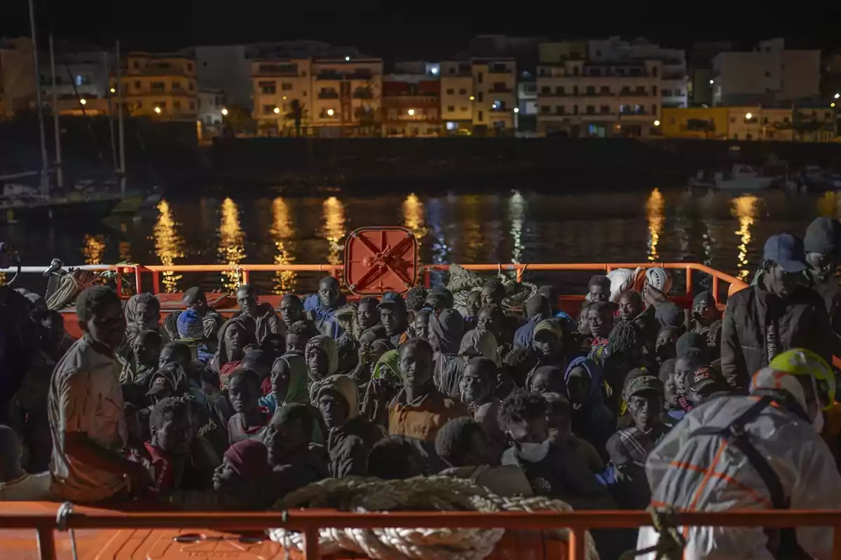 Un grupo de personas se encuentra en un barco de rescate durante la noche, con un fondo de edificios iluminados reflejándose en el agua.