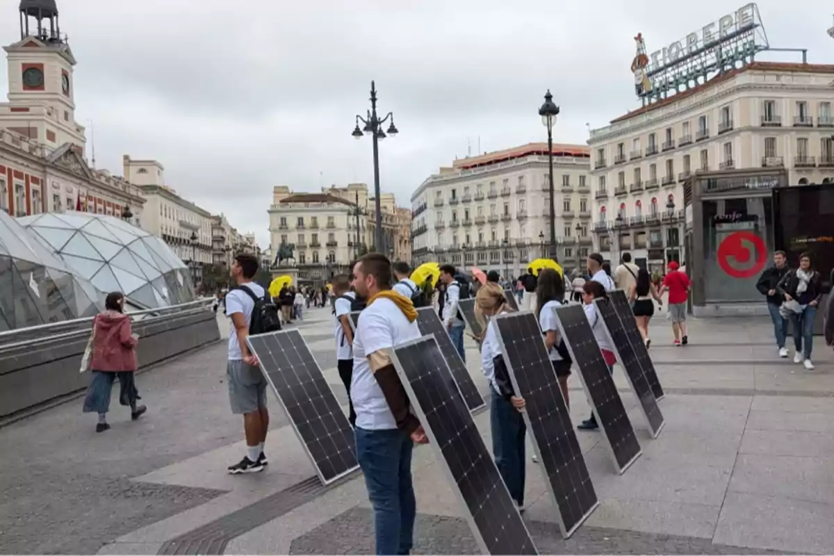 Personas sosteniendo paneles solares en una plaza concurrida con edificios históricos y un reloj en el fondo.