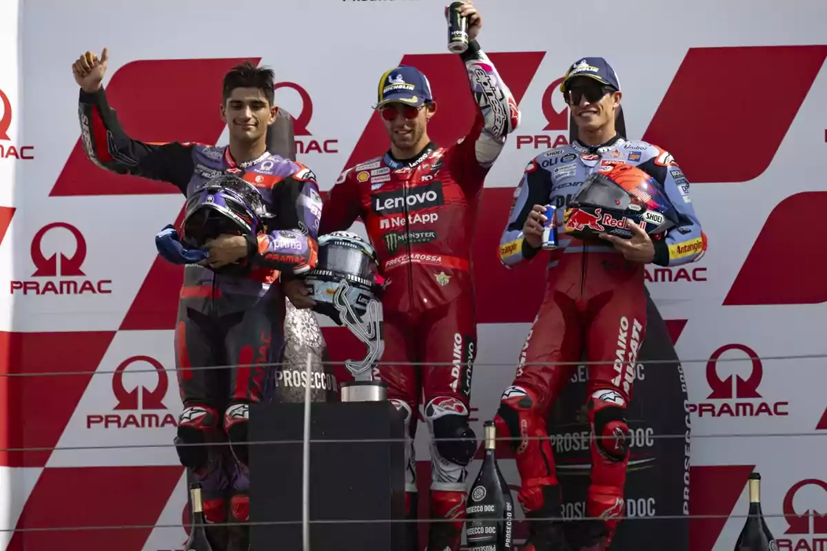 Tres pilotos de motociclismo en el podio de una carrera, celebrando con trofeos y bebidas energéticas, con el logotipo de Pramac en el fondo.