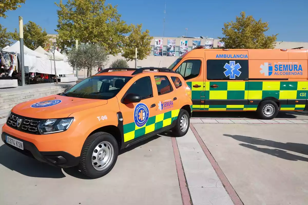 Vehículos de emergencia de SEMURA Alcobendas, incluyendo una ambulancia y un coche de intervención rápida, estacionados en una zona pública con árboles y tiendas de campaña al fondo.