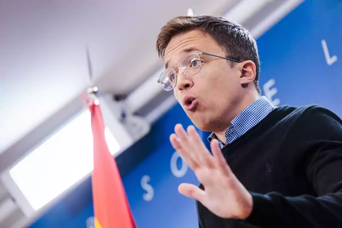 Un hombre con gafas y suéter negro gesticula mientras habla en un entorno con fondo azul y una bandera roja.