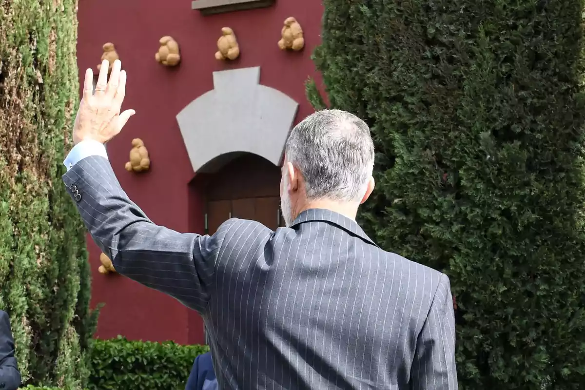 Un hombre de cabello canoso y traje a rayas levanta la mano en señal de saludo, de espaldas a la cámara, con un fondo de árboles y una pared roja decorada con figuras. Un hombre de cabello canoso y traje a rayas levanta la mano en señal de saludo, de espaldas a la cámara, con un fondo de árboles y una pared roja decorada con figuras.