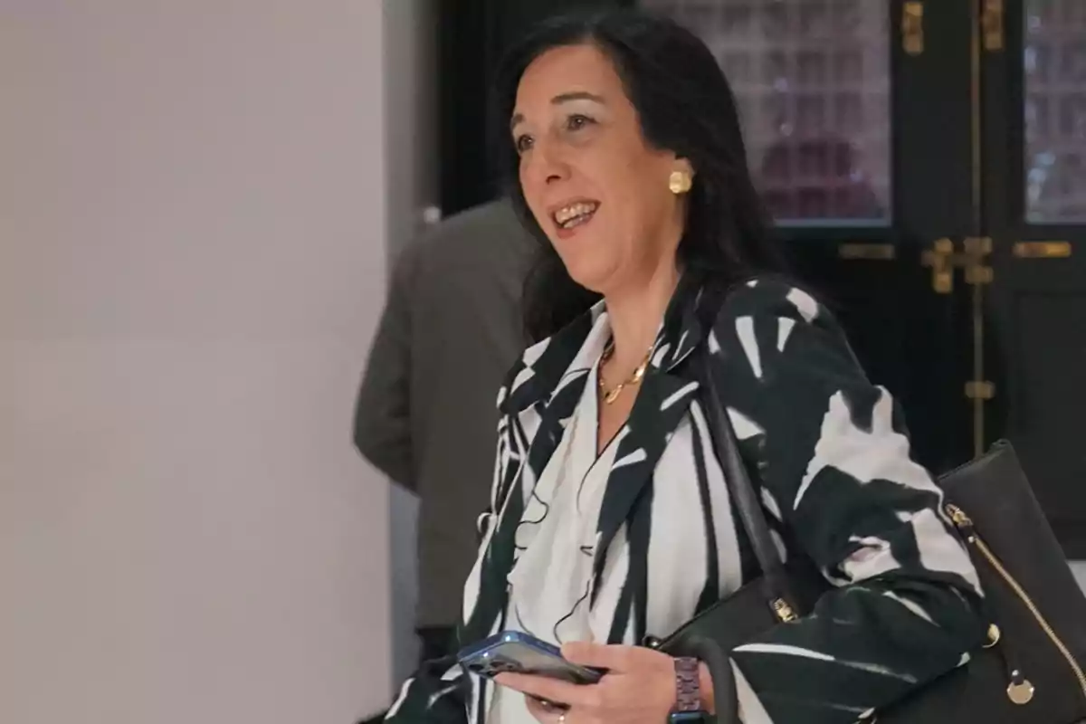 A smiling woman with dark hair carries a black handbag and holds a mobile phone while walking indoors.