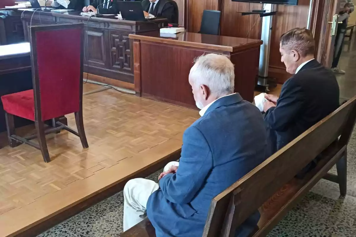 Dos hombres sentados en un banco de madera en una sala de tribunal mientras un juez y abogados están en el estrado. Dos hombres sentados en un banco de madera en una sala de tribunal mientras un juez y abogados están en el estrado.