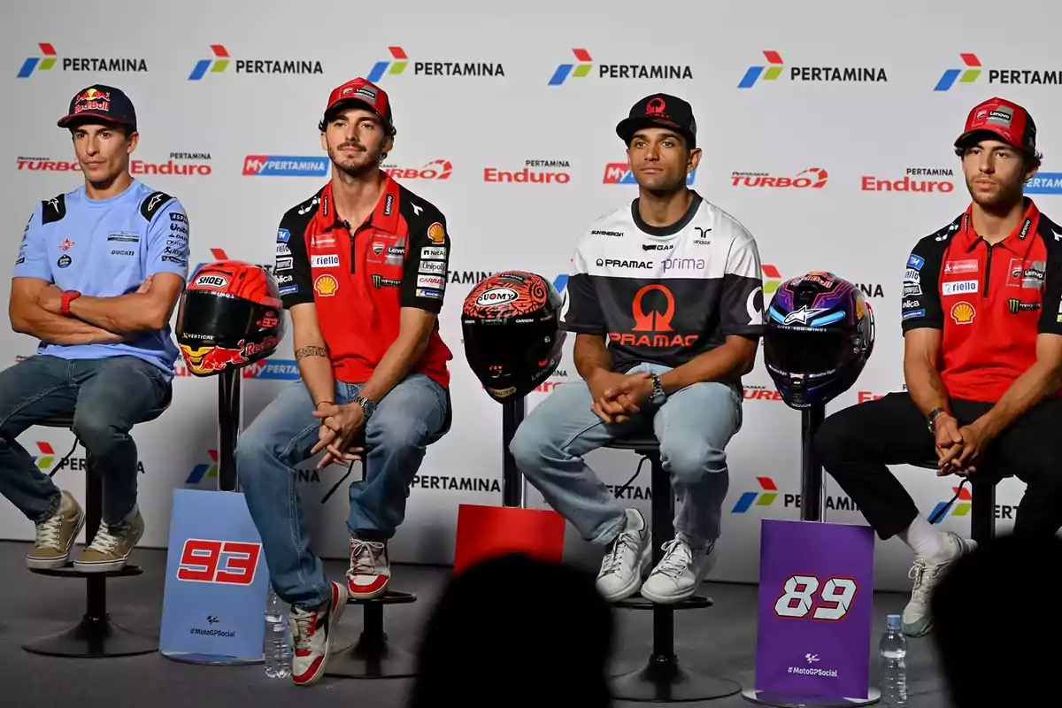 Cuatro pilotos de motociclismo sentados en taburetes con cascos frente a ellos durante una conferencia de prensa, con un fondo que muestra logotipos de patrocinadores.