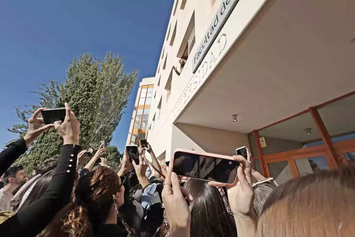 Personas tomando fotos con sus teléfonos móviles frente a un edificio universitario.