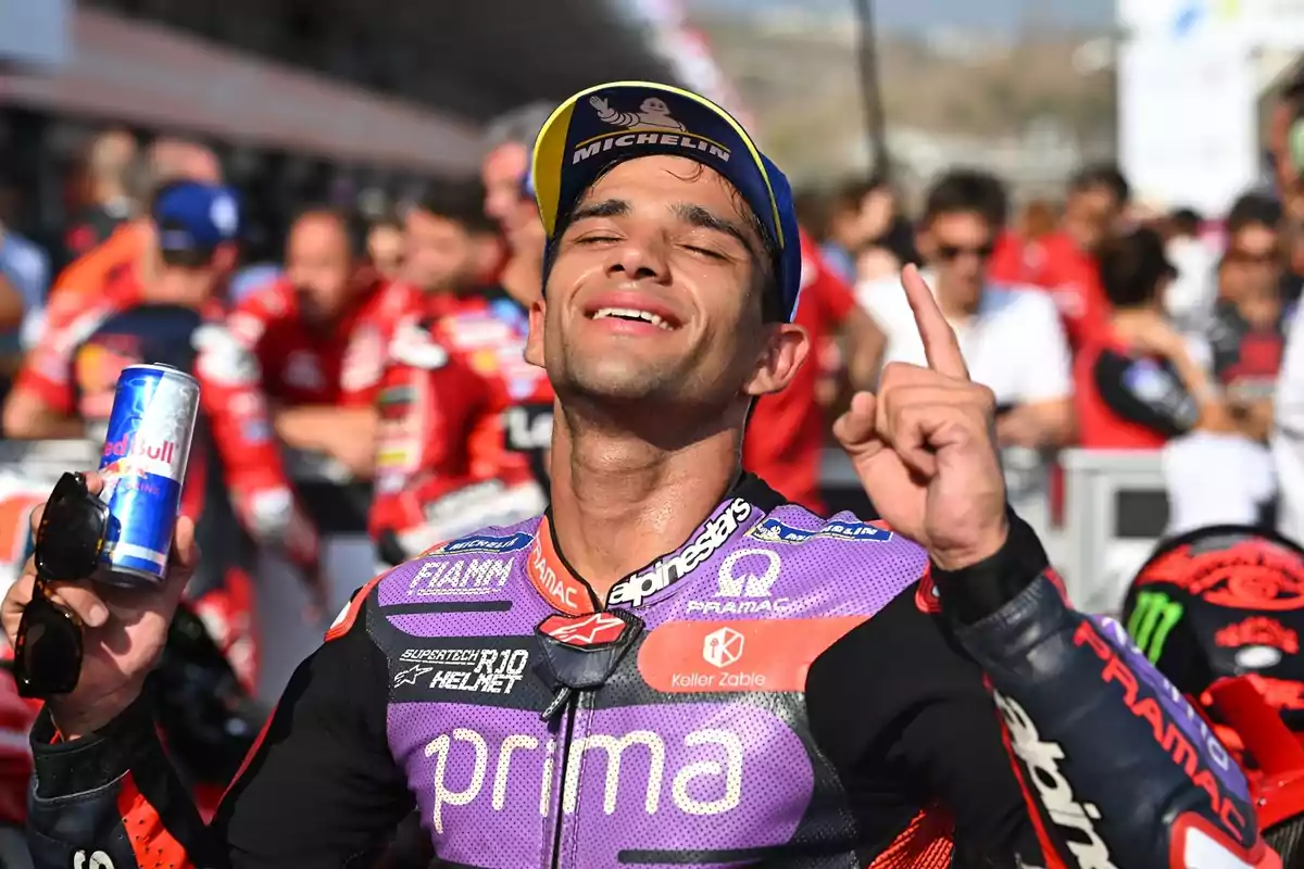 Un motociclista celebrando con una lata de Red Bull en la mano y levantando un dedo, rodeado de personas en el fondo.