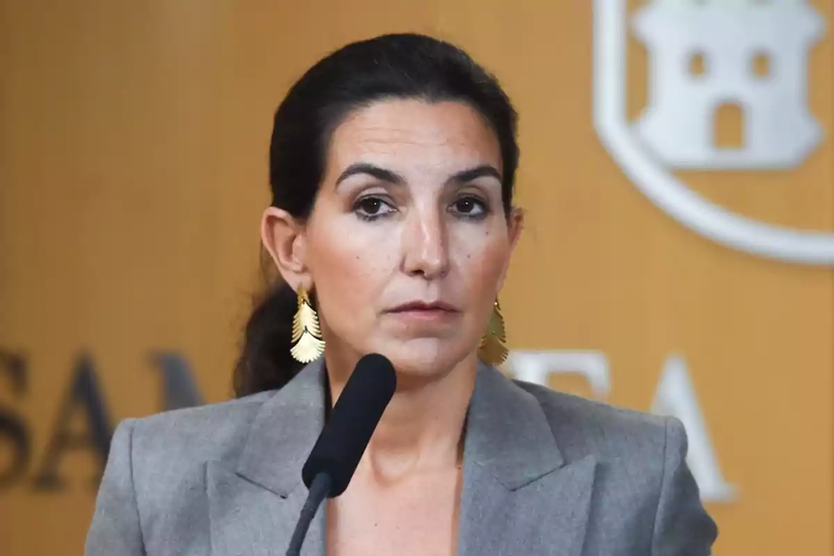 Mujer con cabello oscuro recogido y pendientes dorados frente a un micrófono en un entorno formal.