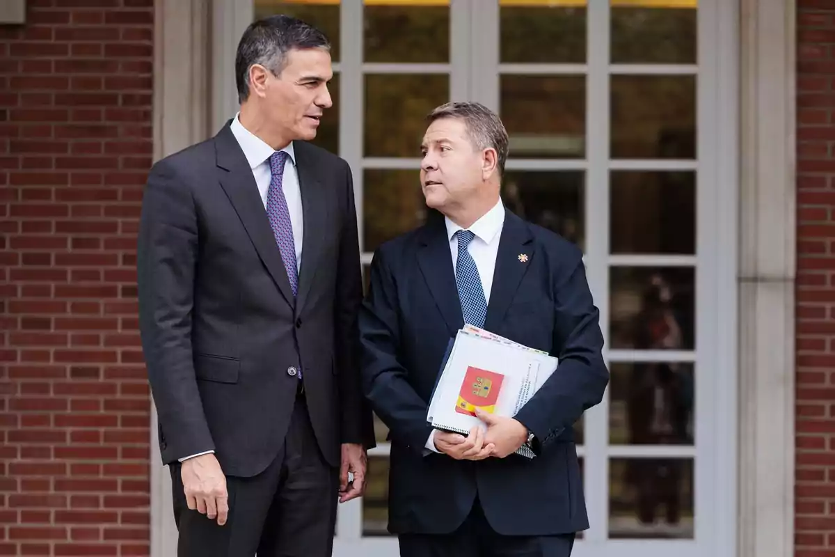 Dos hombres de traje conversan frente a un edificio de ladrillo con puertas de vidrio.