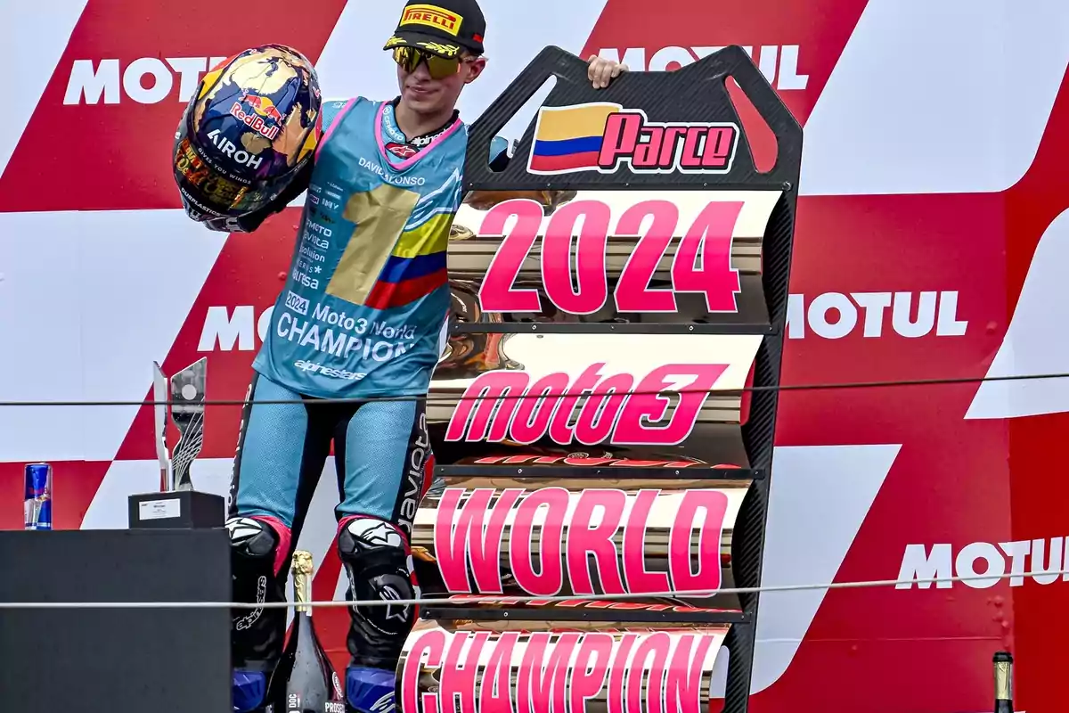 Un piloto de motociclismo celebra en el podio con un cartel que dice "2024 Moto3 World Champion" y sostiene un casco decorado.