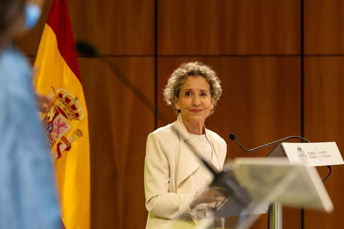 Mujer de cabello corto y canoso hablando en un atril con el escudo de España y una bandera española al fondo en una sala de conferencias Mujer de cabello corto y canoso hablando en un atril con el escudo de España y una bandera española al fondo en una sala de conferencias