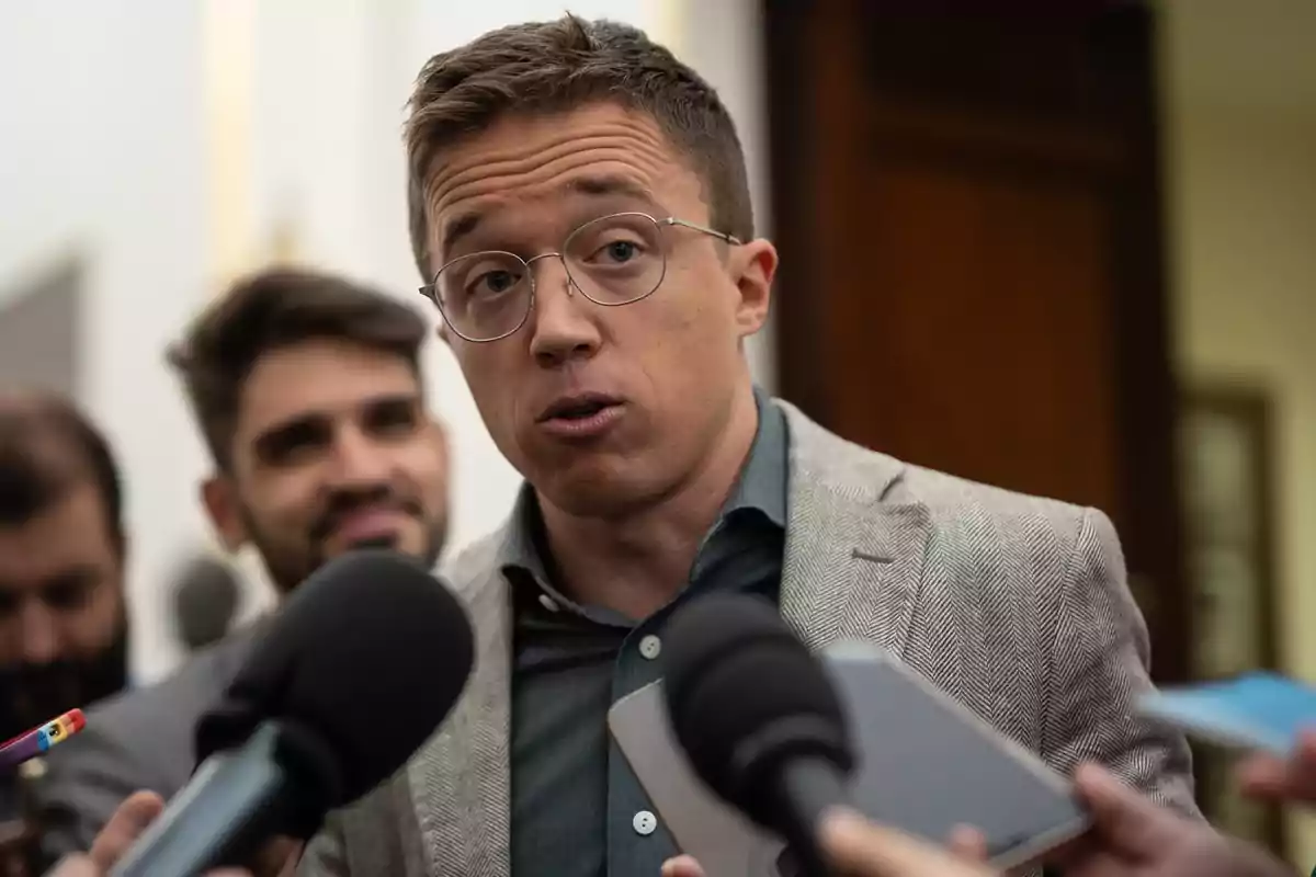 Un hombre con gafas y chaqueta habla frente a varios micrófonos.