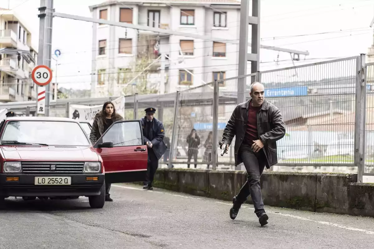 A man with a leather jacket and a gun runs down the street while a woman and a police officer are near a parked red car.