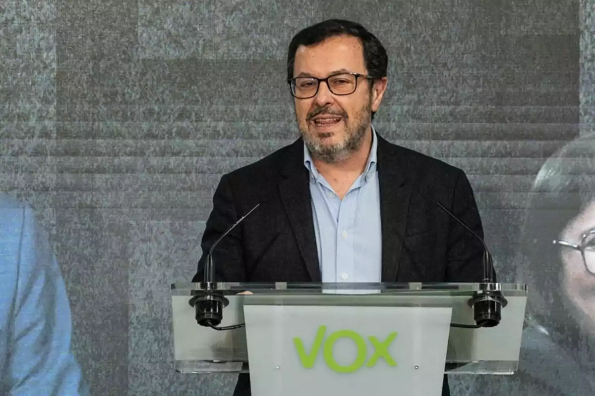 Hombre con gafas y barba hablando en un atril con el logo de VOX