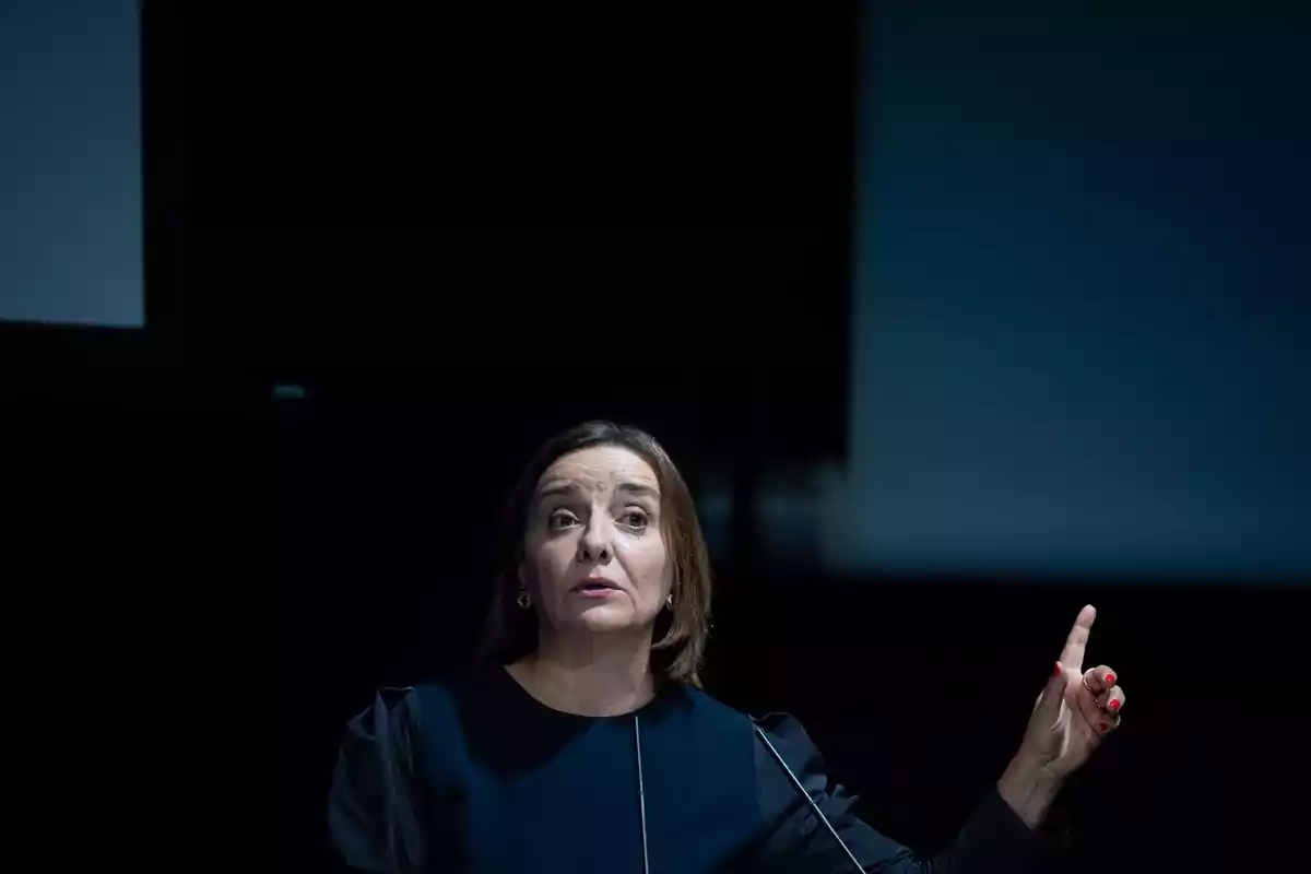 Mujer de cabello castaño y vestido oscuro hablando en público y levantando un dedo mientras gesticula Mujer de cabello castaño y vestido oscuro hablando en público y levantando un dedo mientras gesticula