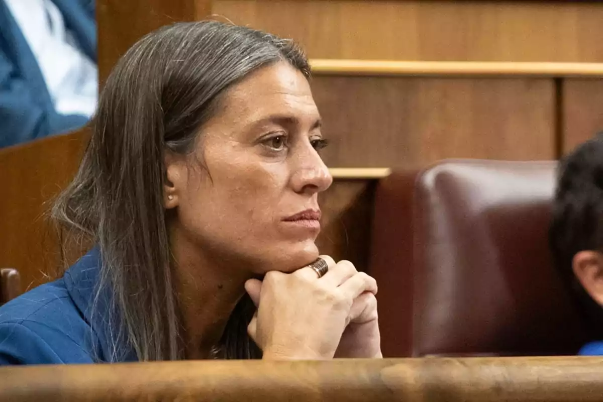 Una persona con cabello largo y oscuro está sentada en un entorno formal con las manos apoyadas en el mentón.