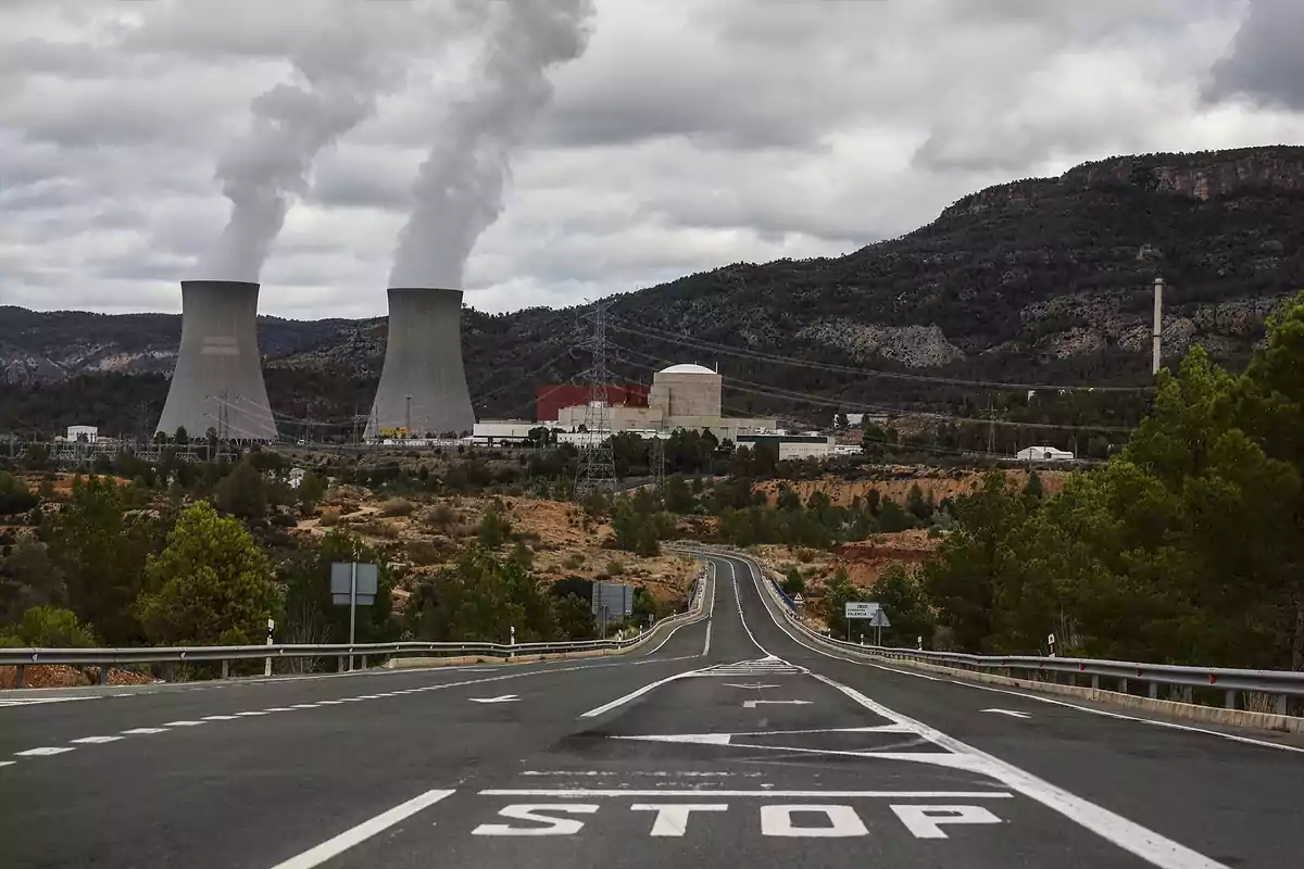 Carretera que conduce a una planta nuclear con dos torres de refrigeración emitiendo vapor en un paisaje montañoso.
