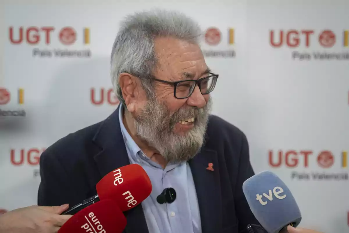 Hombre mayor con barba y gafas sonriendo mientras es entrevistado por varios medios de comunicación frente a un fondo con el logo de UGT País Valencià Hombre mayor con barba y gafas sonriendo mientras es entrevistado por varios medios de comunicación frente a un fondo con el logo de UGT País Valencià