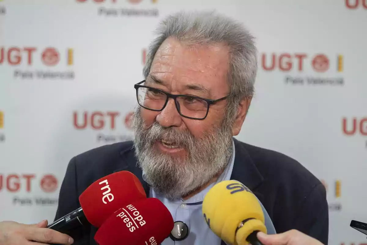 Hombre mayor con barba y gafas hablando ante varios micrófonos de prensa con el logo de UGT de fondo